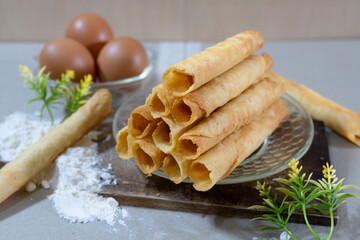 A stack of freshly baked Egg Roll cookies or biscuits displayed with flour and eggs, highlighting homemade baking with simple ingredients. Perfect for a cozy dessert or snack idea.