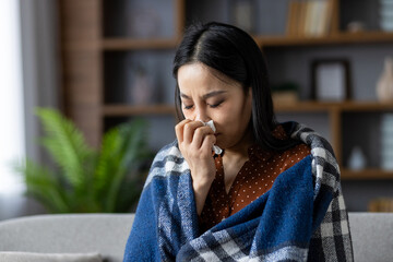 Asian woman sitting at home wrapped in warm blanket while holding tissue. She appears to be unwell, dealing with cold symptoms, seeking comfort and warmth indoors, feeling unwell.