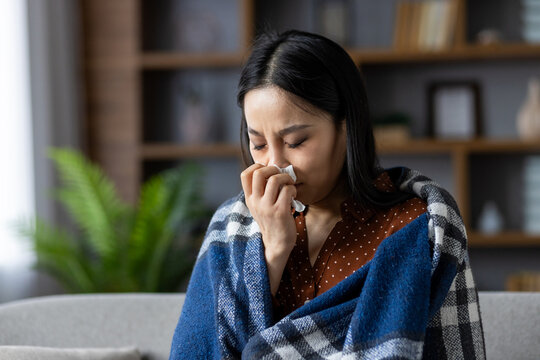 Asian woman sitting at home wrapped in warm blanket while holding tissue. She appears to be unwell, dealing with cold symptoms, seeking comfort and warmth indoors, feeling unwell.