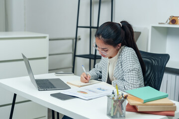 Young Asian woman taking notes while working at desk in an office environment, showing focus and commitment to her tasks, representing a productive and professional workday.
