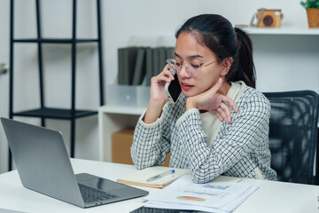 Asian woman holding phone to ear while working at desk with laptop and documents, portraying professionalism, multitasking, and dedication to responsibilities in office setting.