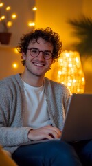 A happy man with glasses is sitting at his laptop in the evening, smiling and looking into the camera