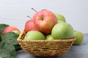 Fresh ripe apples in wicker basket and green leaves on grey table, closeup