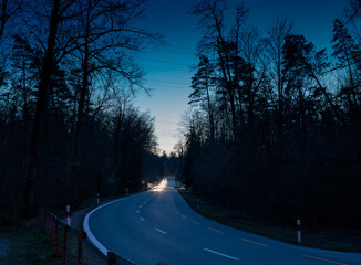 Lonely road in the evening. One single car is driving on the road through the forest. Headlights are producing light trails. © Dirk