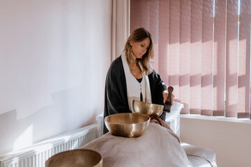 Woman is doing a massage with Tibetan singing bowls in a spa salon against the beige backdrop....