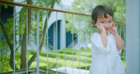 Fototapeta premium A cute Curious child girl in a white dress Exploring enjoys smiling playfully outdoors, standing posing in a lush green garden surrounded by greenery on a sunny day