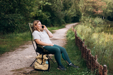Fototapeta premium Relaxed happy woman sitting on deck chair near lake and have conversation on a mobile phone, she is planning new travel. Camping, technology and travel concept. A young 30s woman explores a forest