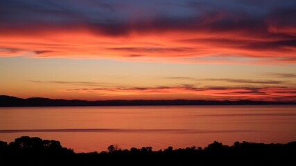 Obraz premium Vibrant orange and pink sunset reflecting on calm water with silhouettes of trees, sky, calm