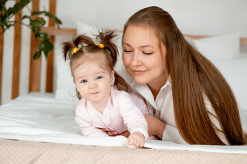 a mother with a baby daughter, a mother gently hugs her baby girl kissing, maternal love and care, a happy family with a baby on the bed at home in the bedroom.