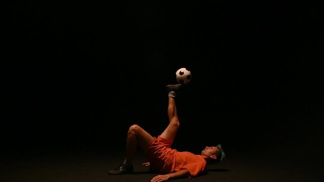 Football freestyle of a guy in an orange uniform on a black background . A football player fills a ball on a black background. A young man juggles a ball. 