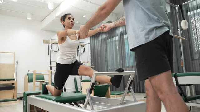 Woman receiving assistance in split stretch exercise during pilates class on trapeze table for flexibility and core training. Student doing split stretch on reformer for improving balance. Habituate.
