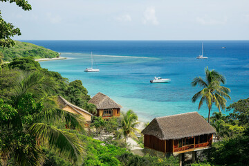 Fototapeta premium Picturesque view of a coastal hill with tropical huts overlooking a turquoise sea. Sailboats are visible in the distance under a clear sky, perfect for a scenic escape.