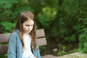 A cute little girl in a sweater sits sadly on a wooden bench in the summer looking at her feet. The concept of children's fun, safety, education, health, insurance