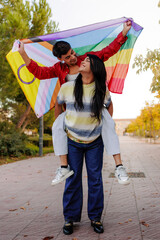 Latin couple holding progress pride flag showing LGBTQIA symbol, piggyback ride in park
