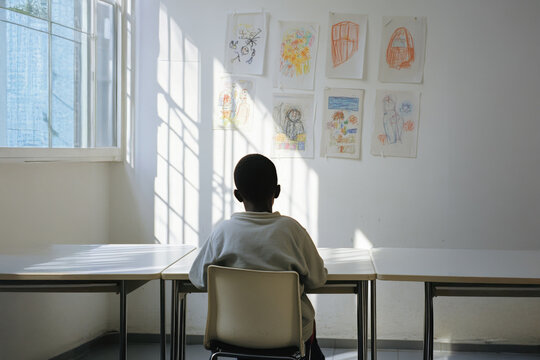 A black child sits alone at a desk in a classroom, gazing thoughtfully at colorful drawings pinned on the wall illuminated by gentle sunlight pouring through the windows. refugee integration