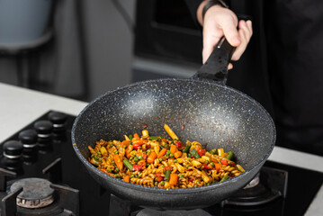 Close up of Chef cook hands cooking and toss roasted pasta, onion, pepper, carrot, vegetables, mushrooms, green peas, beans, garlic in frying wok pan on gas stove. Flying food levitation.