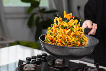 Close up of Chef cook hands cooking and toss roasted pasta, onion, pepper, carrot, vegetables, mushrooms, green peas, beans, garlic in frying wok pan on gas stove. Flying food levitation.
