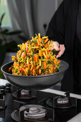 Close up of Chef cook hands cooking and toss roasted pasta, onion, pepper, carrot, vegetables, mushrooms, green peas, beans, garlic in frying wok pan on gas stove. Flying food levitation.