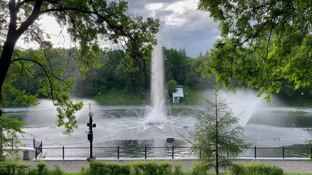 Beautiful fountain spraying water in city pond in summer with green trees around