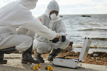 Side view closeup of two people wearing protective suits taking samples and probes in nature outdoors sitting by water and using toolkit, copy space