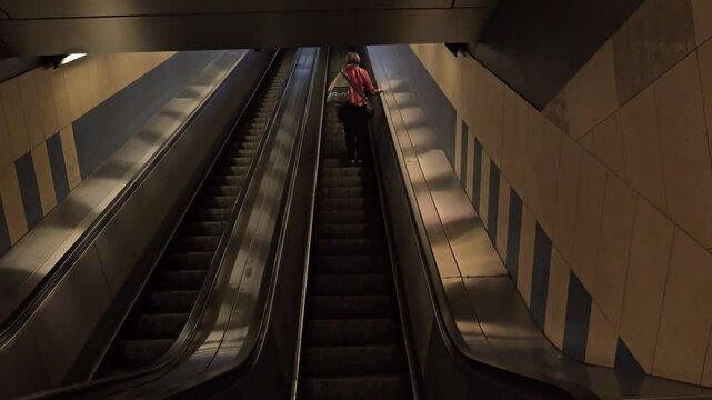 A solitary woman ascends an escalator in a dimly lit metro station. The setting evokes feelings of solitude and urban exploration, highlighting geometric patterns on the walls