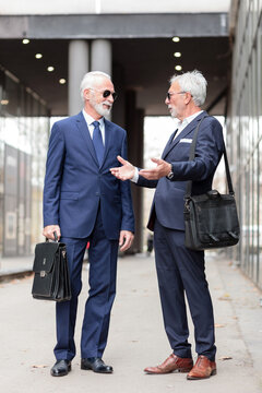 Two happy gray haired senior Caucasian businessmen standing on the sidewalk in front of office building and talking
