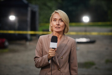 Waist up portrait of blonde adult woman as female reporter speaking to microphone while covering...