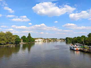 London, UK - 05.09.2024: View of Tideway of River Thames with a boat moored at Kew Pier on the right and trees and houses along the riverbank under a clear blue sky with clouds, view from Kew Bridge