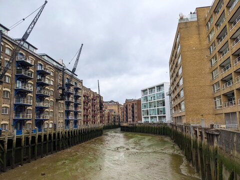 London, UK - 05.13.2024: A row of historic dockside buildings and warehouses with cranes in New Concordia Wharf overlooking the exposed mudflats of the River Thames at low tide under a cloudy sky