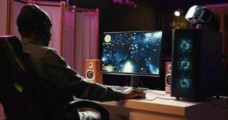 Man coming home to play SF space videogame on PC screen after finishing job shift. Gamer entering room, starting spaceship flying game on computer in RGB lit home late at night, camera B