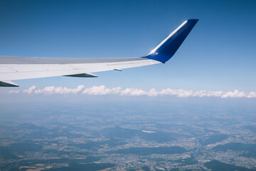 Obraz premium View from an airplane window of a wing against a clear blue sky, with clouds and a scenic landscape below. Captures the essence of air travel and aerial perspective.
