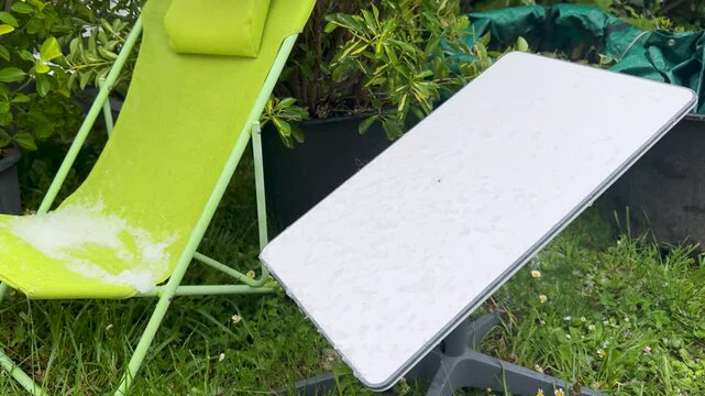 A satellite dish stands in a garden alongside a green deck chair covered with hail, capturing the scene of a recent hailstorm - outdoor technology enduring extreme weather.
