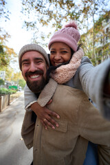 Fototapeta premium Vertical. Winter portrait of happy young tourist couple taking piggyback selfie outdoors in a city. Smiling multi-ethnic people traveling together on vacation. Cheerful people posing for photo phone