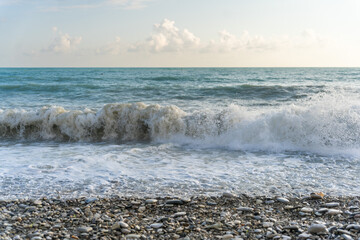 The sea waves roll onto a pebble sea shore against a blue sky with clouds.