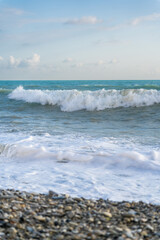 The sea waves roll onto a pebble sea shore against a blue sky with clouds.