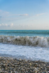 The sea waves roll onto a pebble sea shore against a blue sky with clouds.