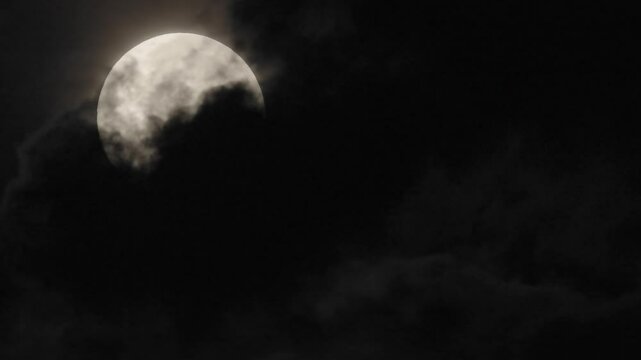 4k video of clouds drifting past the full harvest moon in a dark night sky with the moon partly visible one moment and fully visible the next as it slowly sinks towards the western horizon. 