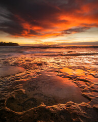 The coastline glows with intense hues as the sun sets over the waters of Sydney, casting a warm,...