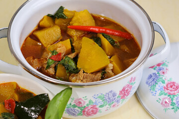 Pumpkin curry with pork, red chillies and basil leaves on top in a glazed pot on a cream-colored marble table.

