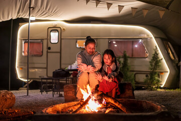 Asian family mother and child camper or kid girl travel camping sitting together on log for warming themselves by bonfire at night with campervan or RV motorhome in holiday at Doi Bo Luang Forest Park