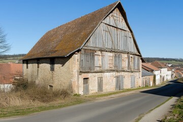 Obraz premium A rustic, weathered barn stands beside a quiet road in a rural landscape.