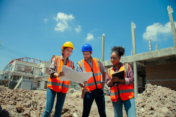 Three construction workers are standing in a field, looking at a blueprint. They are wearing orange...