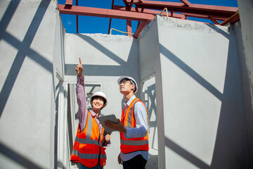 Two construction workers are standing in front of a building, one pointing to something on the other's tablet