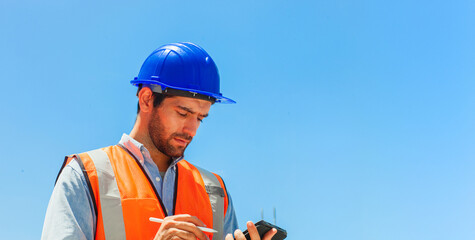 A man in a blue hard hat and orange vest is checking his phone. He is wearing a safety vest and a...