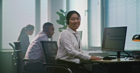 Asian female technical support operator wearing headset, sits in front of computer, looks at camera providing customer service. Team of diverse helpdesk specialists work in modern call center office.