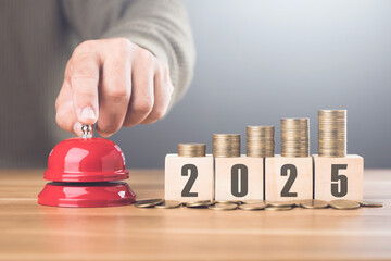 Hand presses a red bell next to a stack of coins on a wooden block labeled '2025,' representing financial goals and future planning with a sense of urgency for the upcoming year