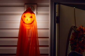 A spooky Halloween decoration hangs by a doorway, casting an eerie glow. A pumpkin lantern, partially obscured by an orange fabric, creates a mysterious and haunting silhouette.