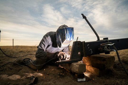 A man in a welding helmet welds a trailer hitch on a farm.