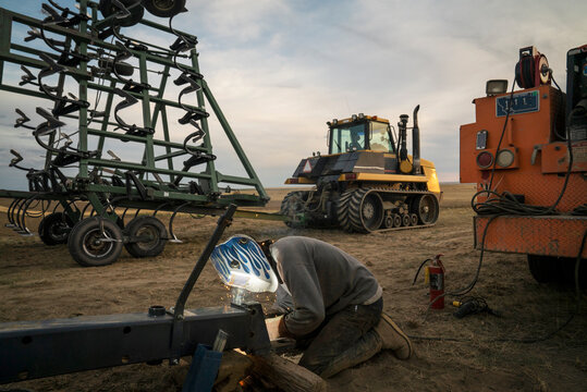 A man in a welding helmet welds a trailer hitch on a farm.