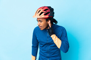 Young cyclist man with braids over isolated background listening to something by putting hand on the ear